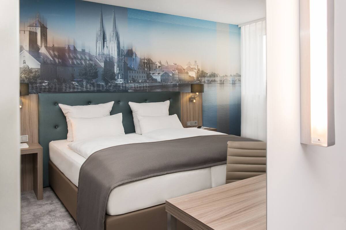 Une chambre d’hôtel avec un lit, des tables de chevet, un bureau et une vue sur le paysage urbain par la fenêtre.