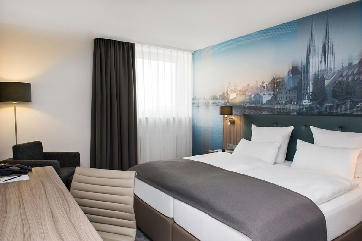 Habitación de hotel con una cama grande, un escritorio, una silla, una lámpara y un mural con vista a la ciudad en la pared.