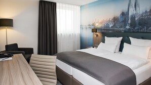 Habitación de hotel con una cama grande, un escritorio, una silla, una lámpara y un mural con vistas a la ciudad en la pared.