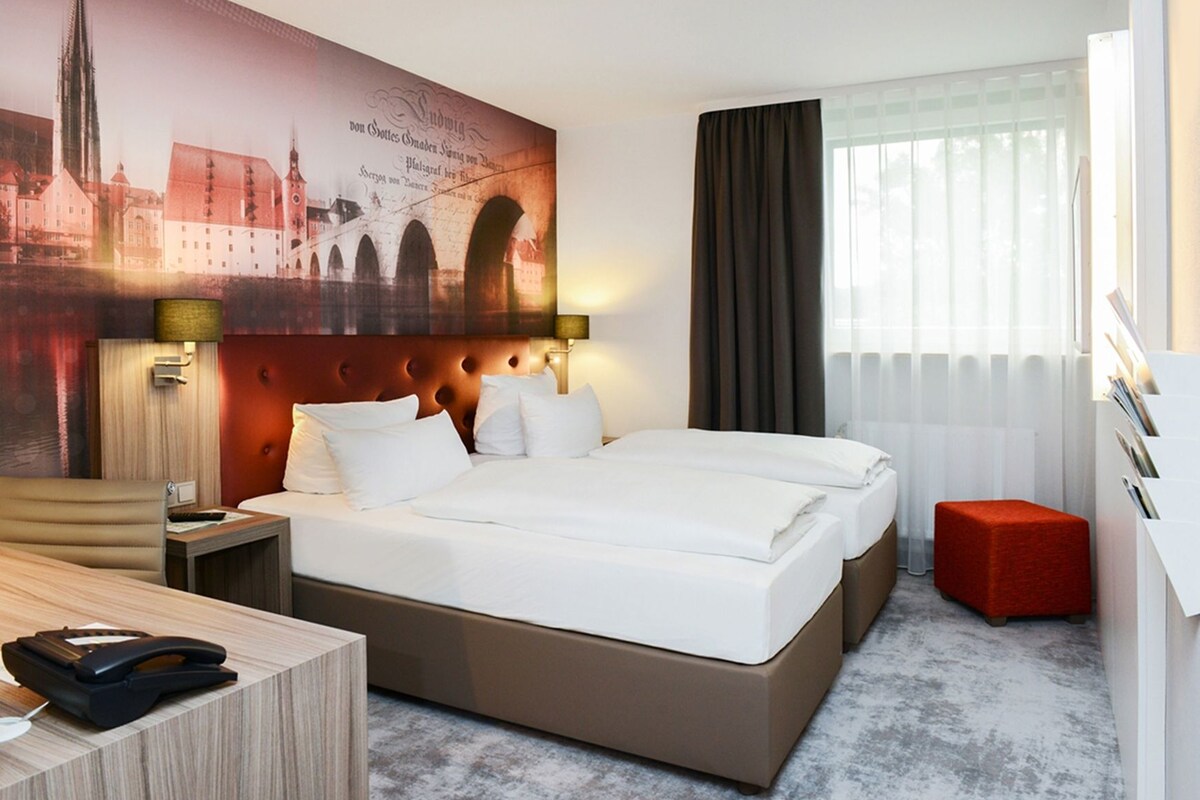 Habitación de hotel con una cama grande, un escritorio, un teléfono, un reposapiés rojo y un mural con vistas a la ciudad.