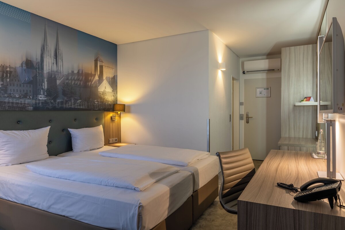 A hotel room with a large bed, a desk with a telephone, and a cityscape painting on the wall.