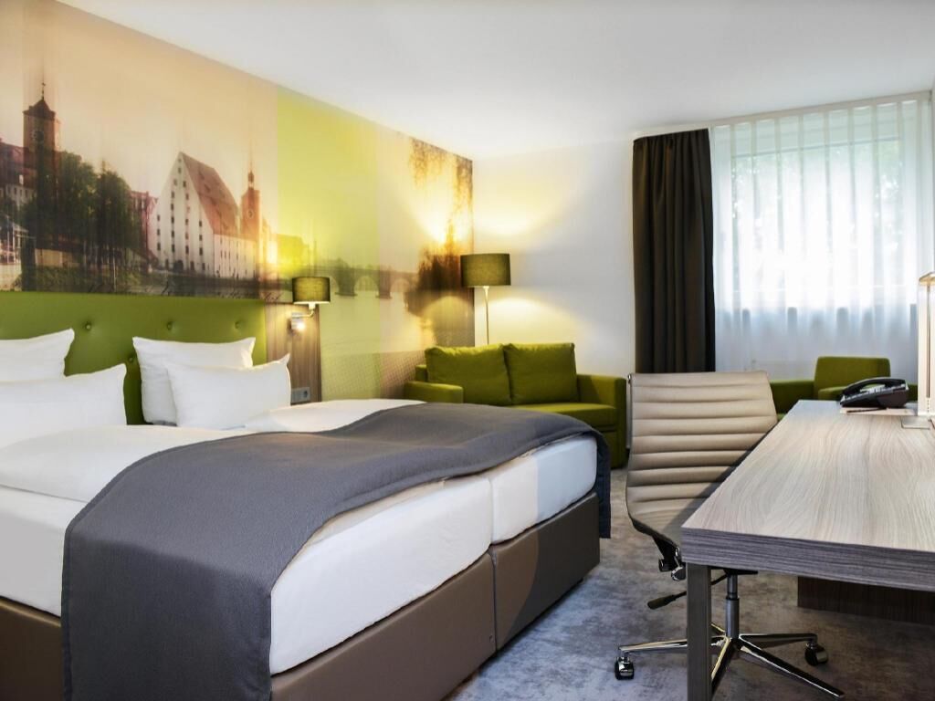 A hotel room with a bed, a desk, a chair, a lamp, and a cityscape painting on the wall.