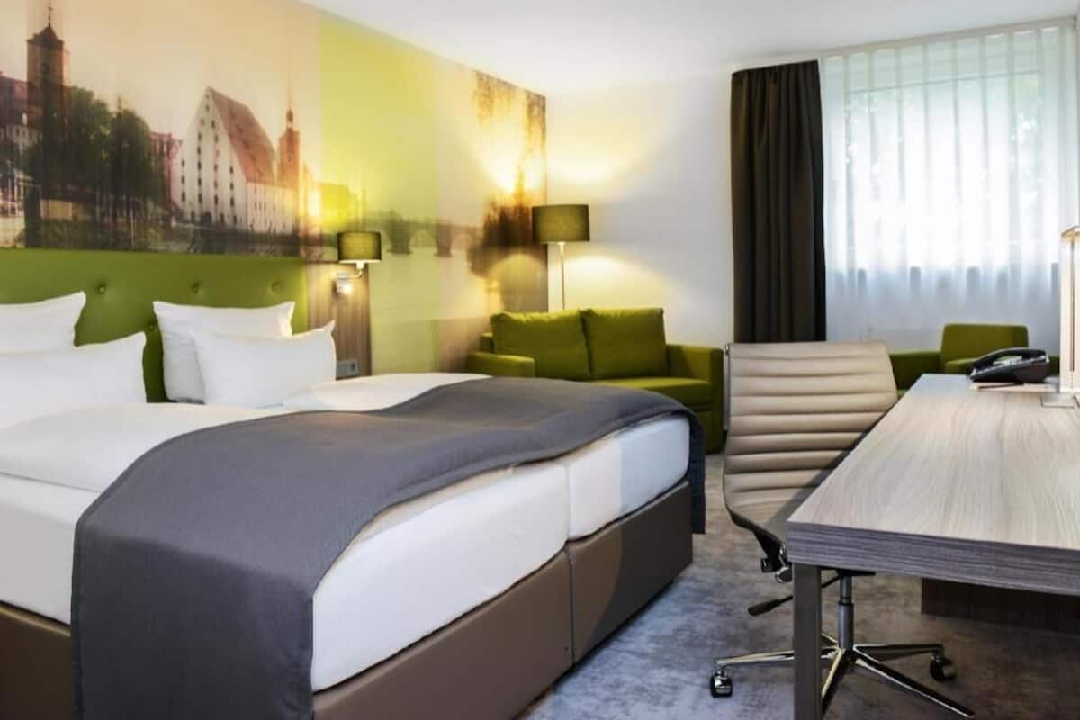 A hotel room with a bed, a desk, a chair, a lamp, and a cityscape painting on the wall.