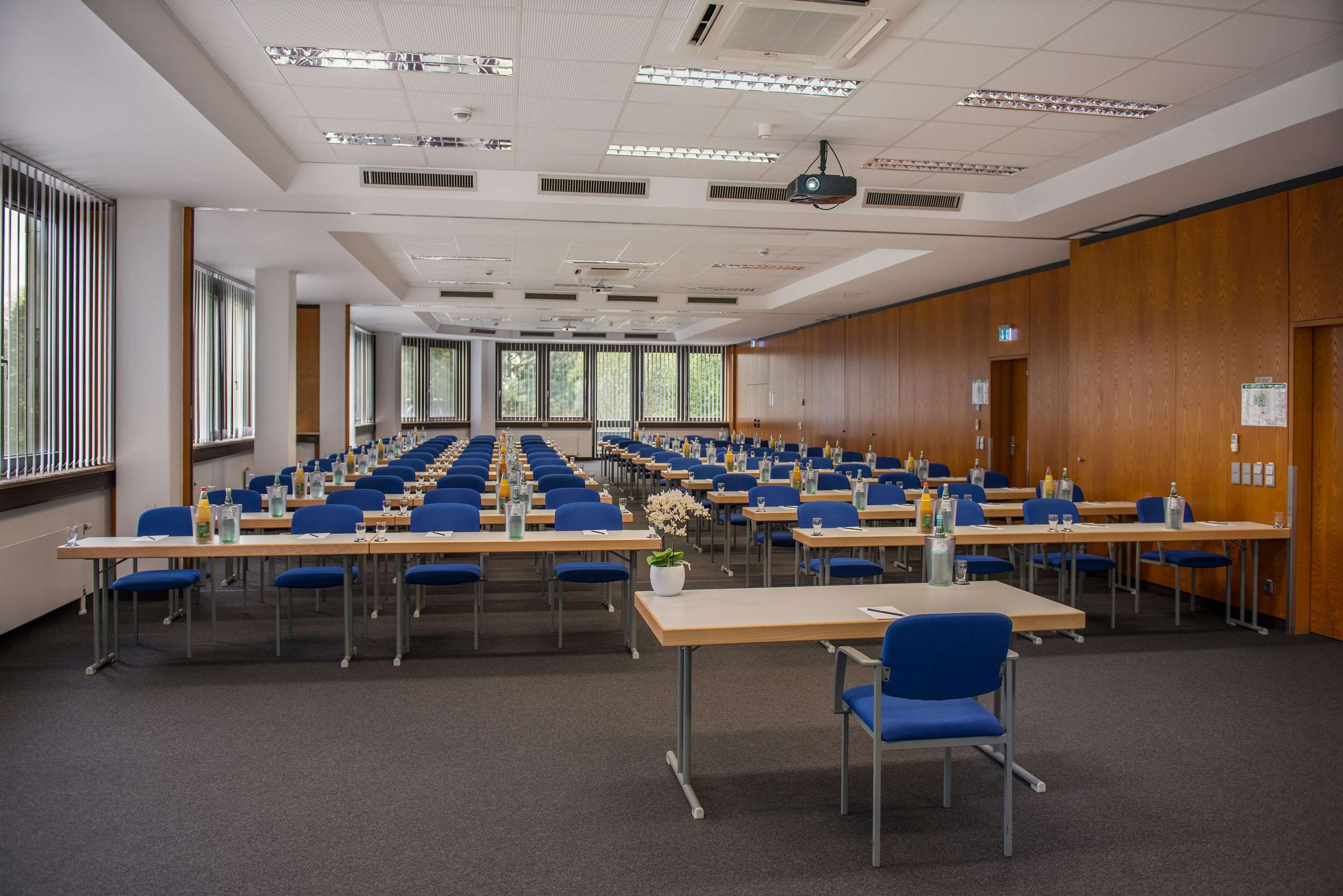 A conference room with rows of tables and chairs, large windows, and a projector on the ceiling.