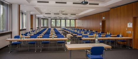 A conference room with rows of tables and chairs, large windows, and a projector on the ceiling.