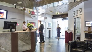 A hotel lobby with a reception desk, a digital screen, a clock, and a glass partition. - Hotel Berlin (Sindelfingen)