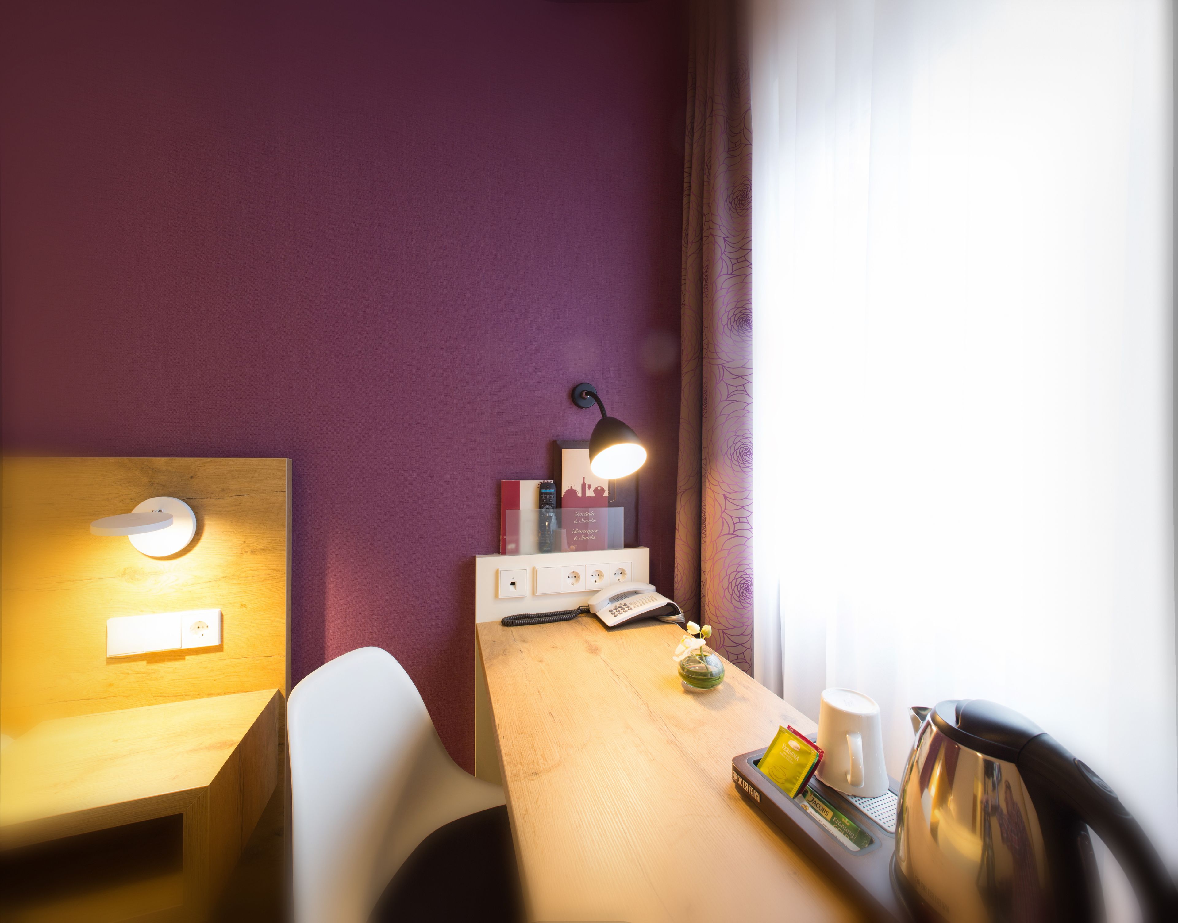 Ein Hotelzimmer mit Schreibtisch aus Holz, einem weißen Stuhl, einem kleinen Tisch mit Lampe, einem Telefon, einer Kaffeemaschine und einem Tablett mit Teebeutel und Tasse.