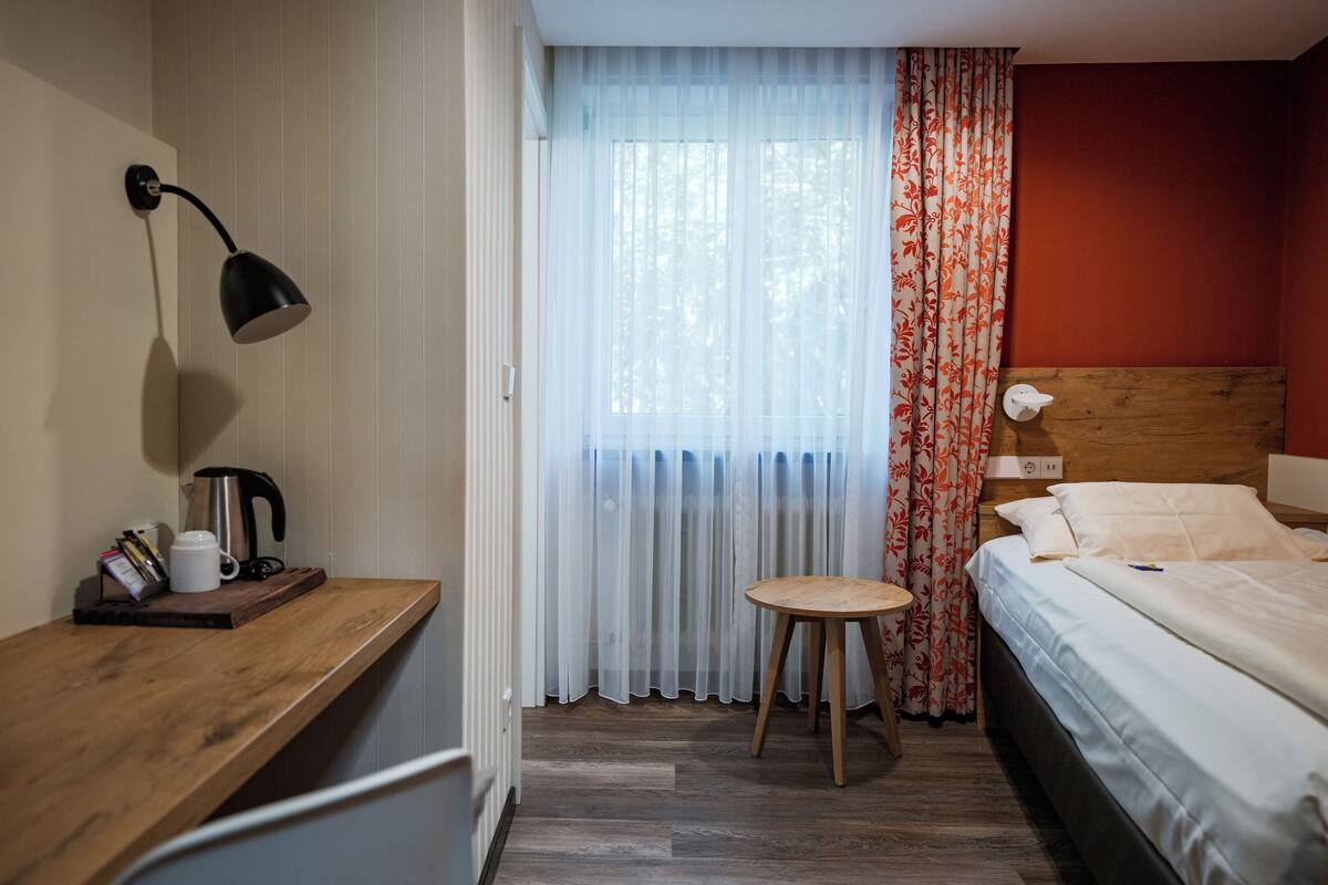Une chambre d’hôtel avec un bureau en bois, un lit, une table de chevet, une lampe et une fenêtre ornée de rideaux légers et d’un voilage à motifs floraux.