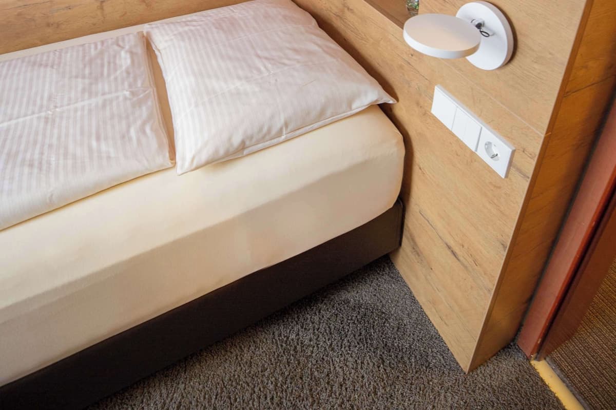 Une petite chambre avec un lit simple, une tête de lit en bois, une table de chevet avec un miroir et un mur blanc.