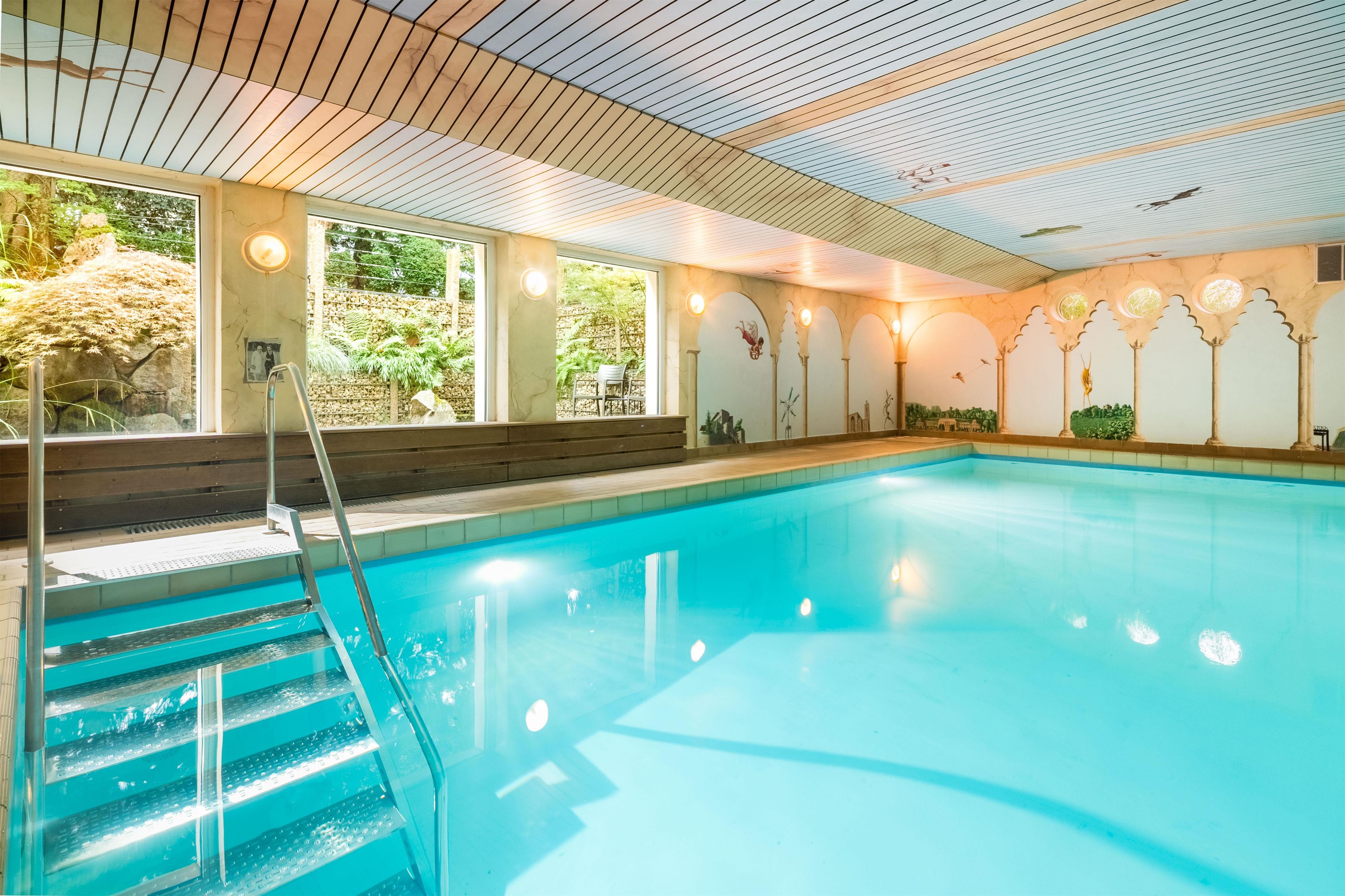 An indoor swimming pool with clear water, a wooden deck, and a view of greenery outside.