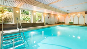 An indoor swimming pool with clear water, a wooden deck, and a view of greenery outside.