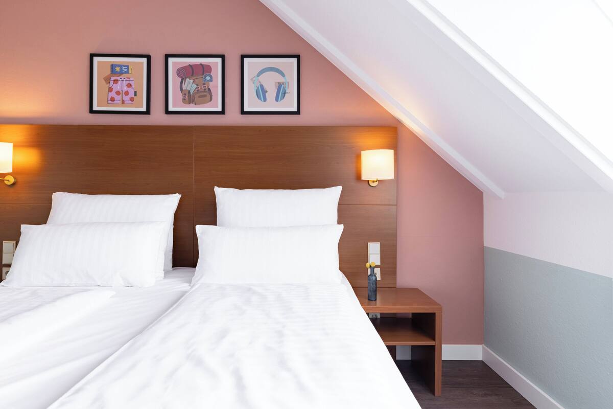 Habitación de hotel con cabecera de madera, dos camas blancas, una mesita de noche con lámpara y tres cuadros en la pared.