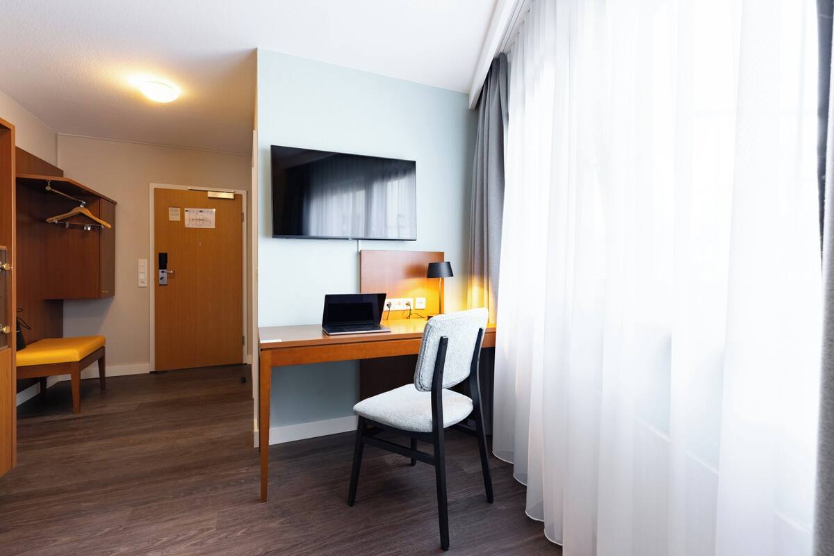 A hotel room with a wooden desk, a laptop, a chair, a TV mounted on the wall, and a door with a sign.