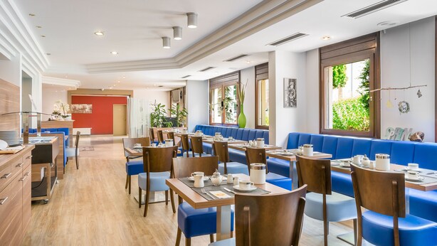 Een moderne eetruimte met blauwe zitbanken en houten tafels, met een lichte en ruimtelijke sfeer dankzij grote ramen.