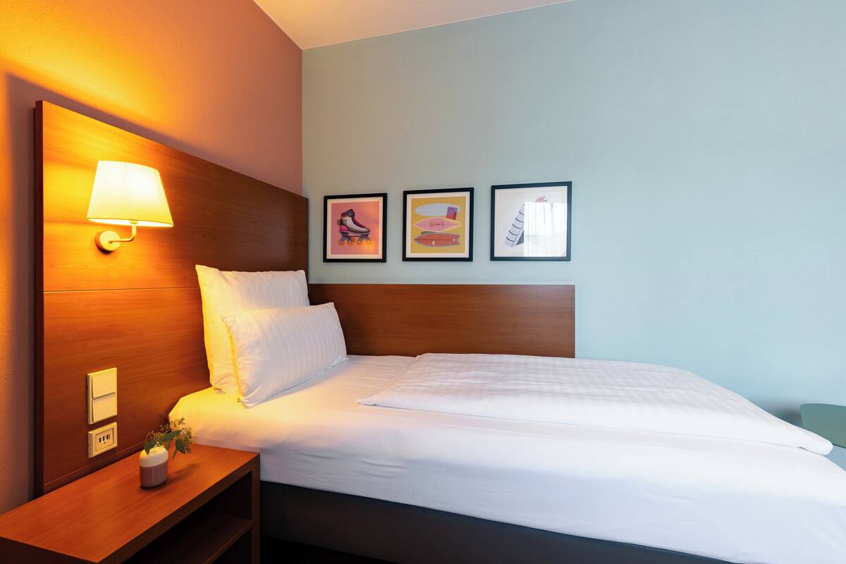 Habitación de hotel con cabecera de madera, lámpara de noche, mesita de noche con un florero y tres cuadros en la pared.