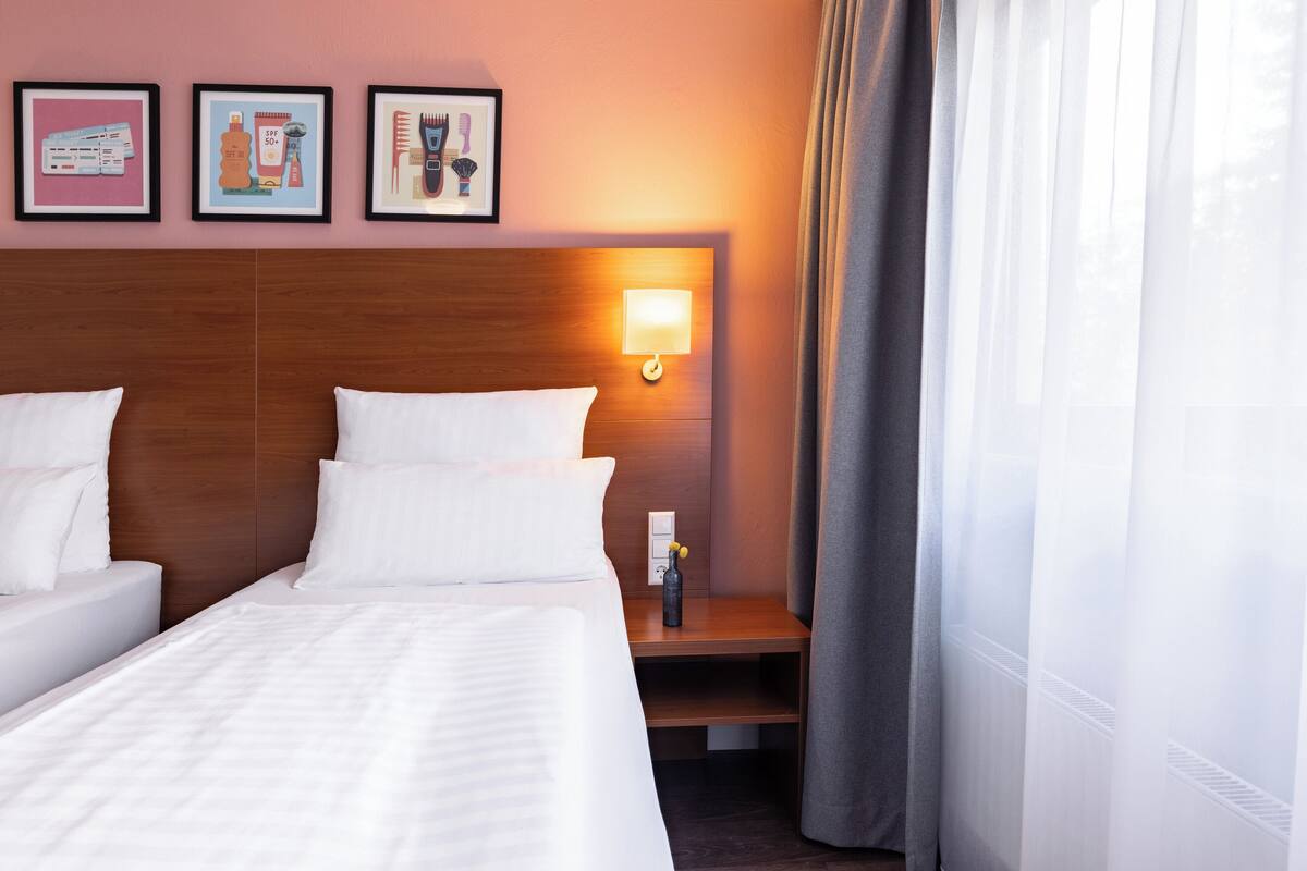 Una habitación de hotel con cabecera de madera, cama con ropa de cama blanca, mesita de noche con lámpara y ventana con cortinas traslúcidas.