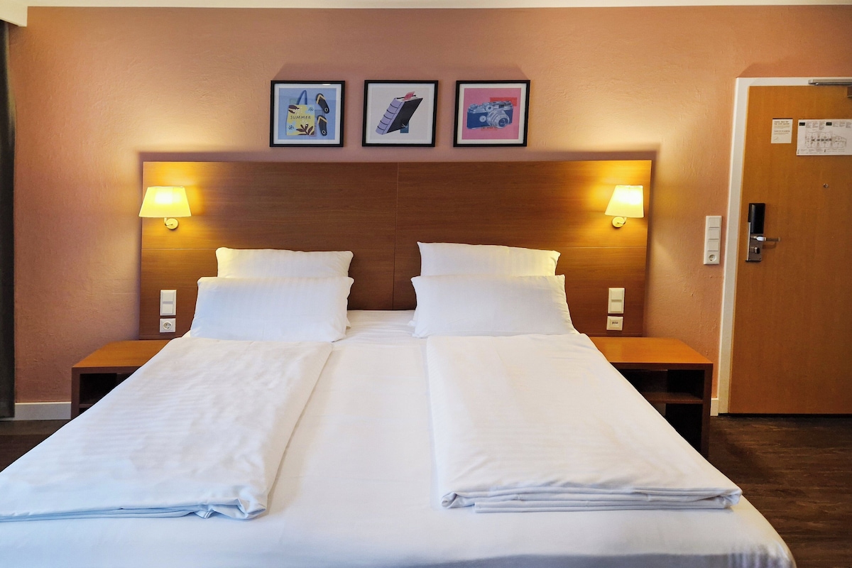 A double bed with white linens, two bedside tables with lamps, and three framed pictures on the wall.