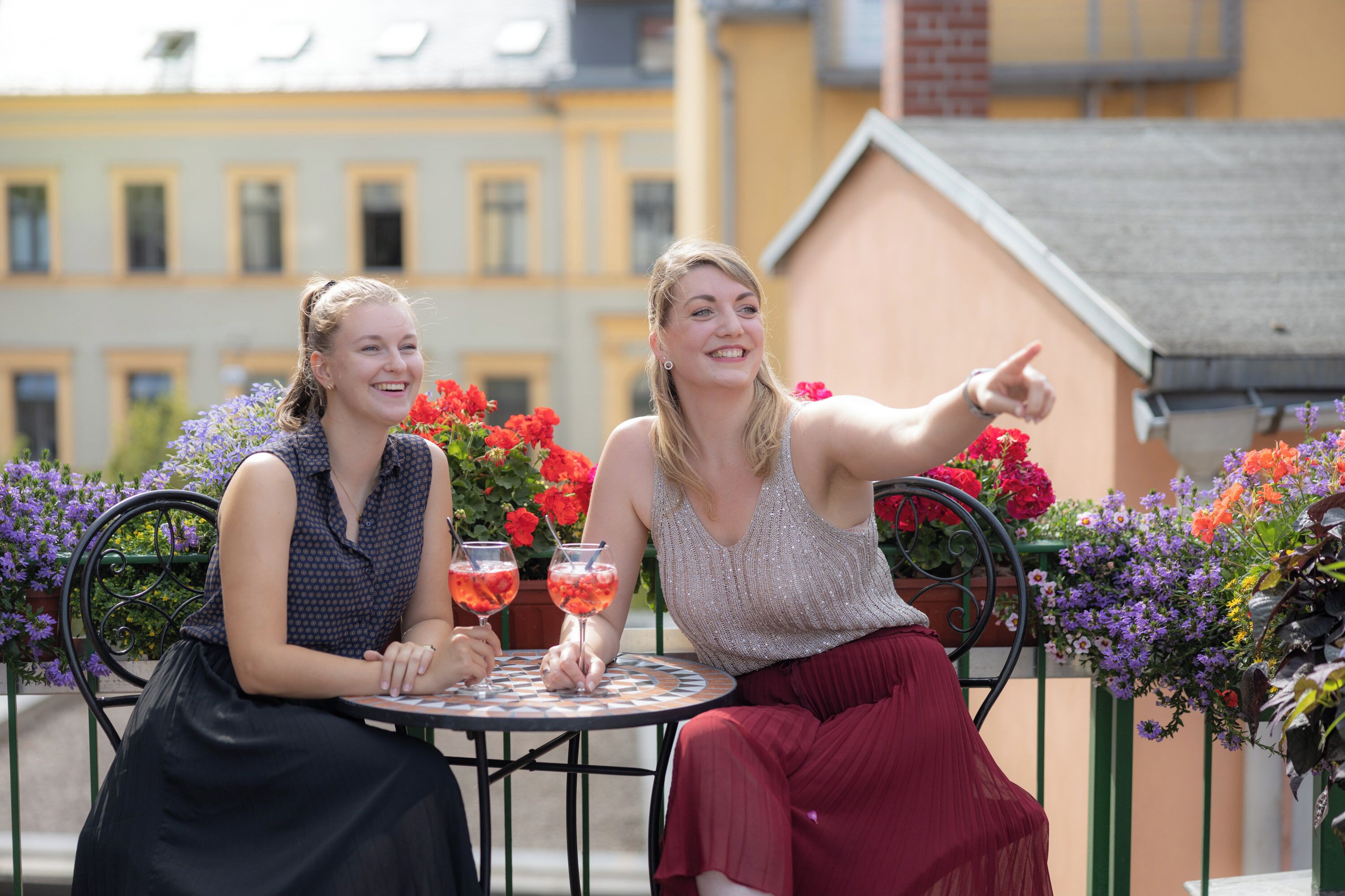 Two people sitting at a table with drinks, surrounded by potted plants and flowers.