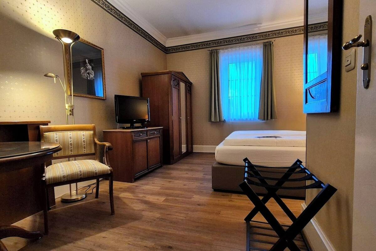 Habitación de hotel con escritorio de madera, una silla, televisor, armario, cama y ventana con cortinas.