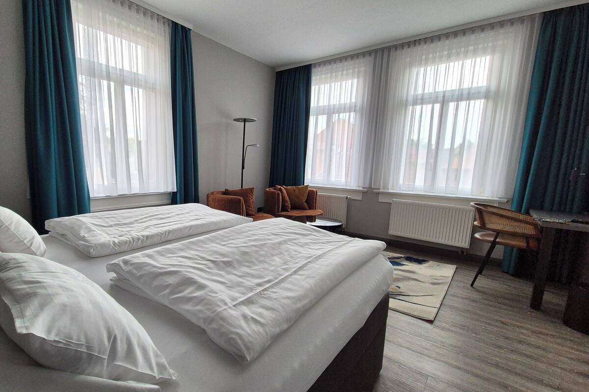 Habitación de hotel con dos camas, un escritorio, una silla y ventanas amplias con cortinas.
