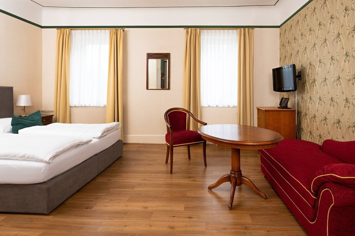 Habitación de hotel con cama, sofá rojo, mesa redonda, televisor y dos ventanas con cortinas.
