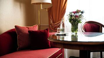 A hotel room with a red sofa, a round glass table, a vase with flowers, a lamp, and a window with curtains.