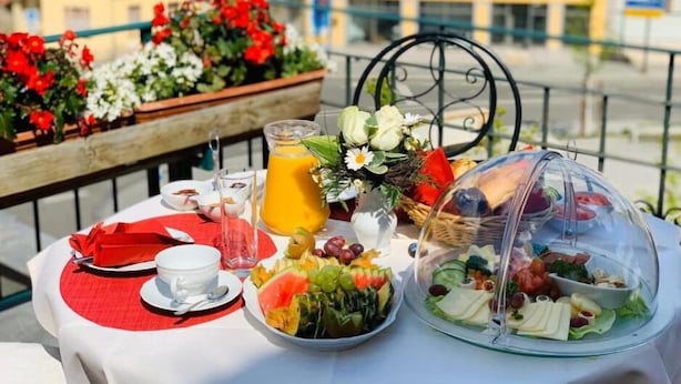 Een gedekte tafel met ontbijtartikelen, waaronder een glas sinaasappelsap, een bord fruit en een afgedekte schaal met diverse snacks, met uitzicht op een balkon.