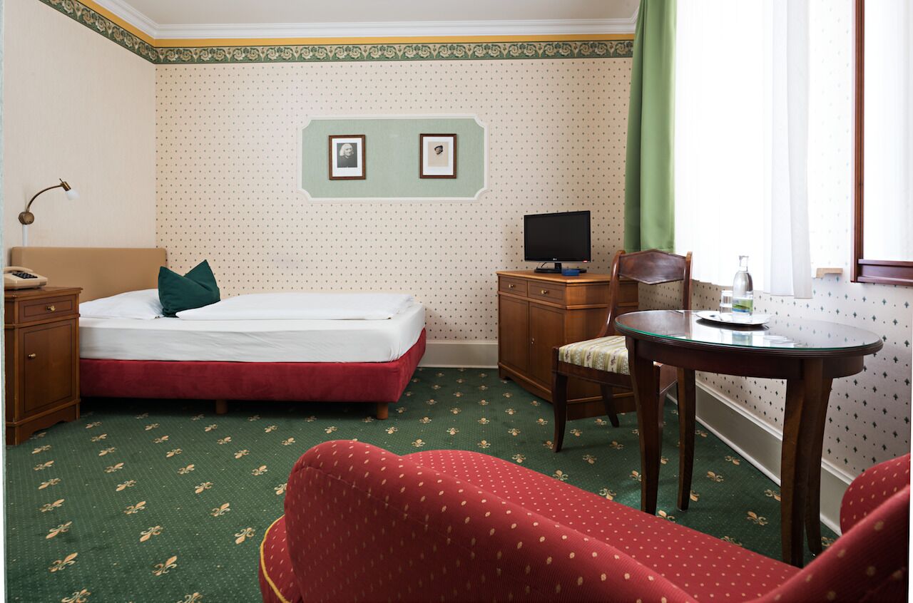 A hotel room with a bed, a desk with a computer, a chair, a small table, and a window with curtains.