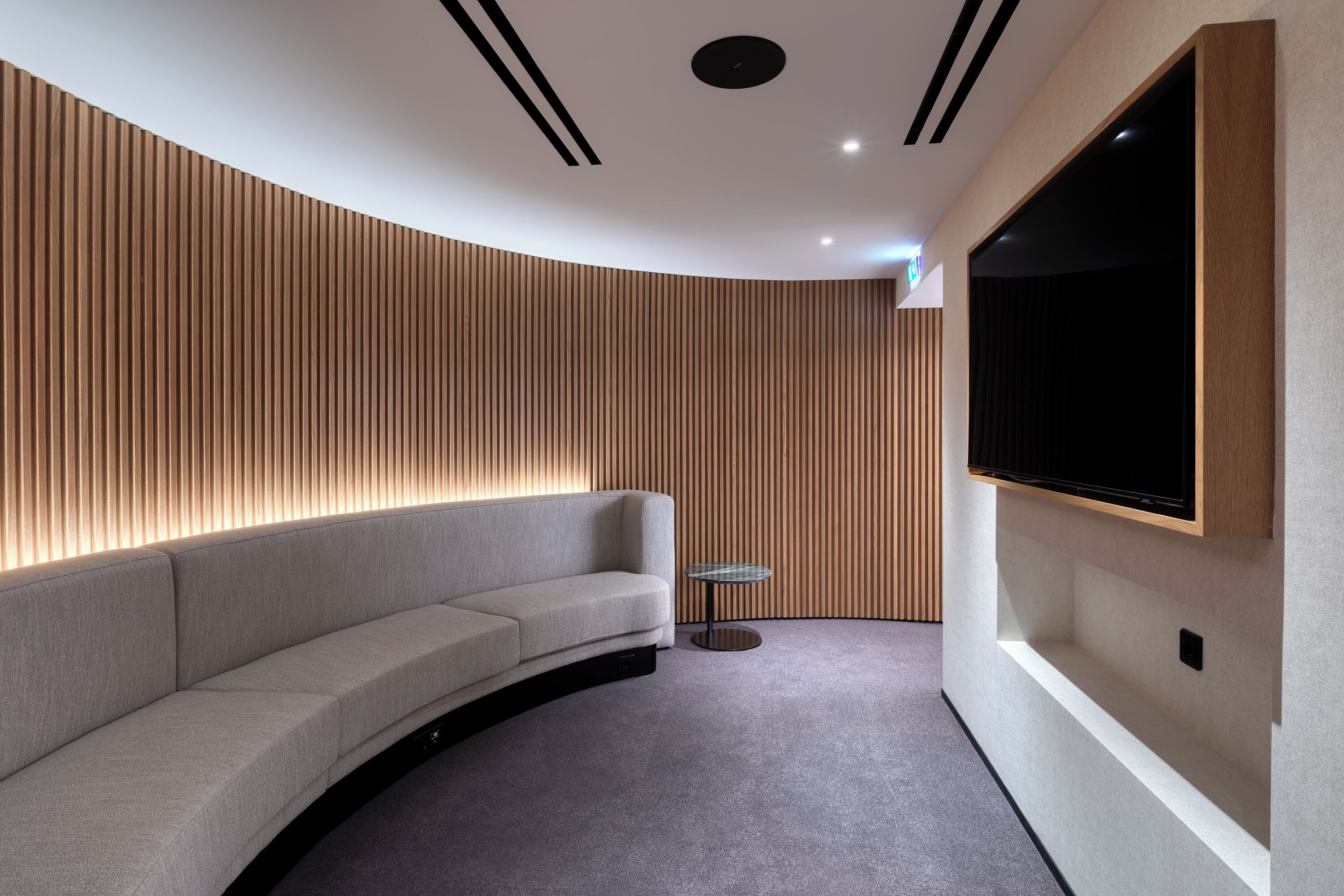 A modern, curved sofa with a built-in TV shelf and a small round table in a contemporary room with wooden paneling and recessed ceiling lighting.
