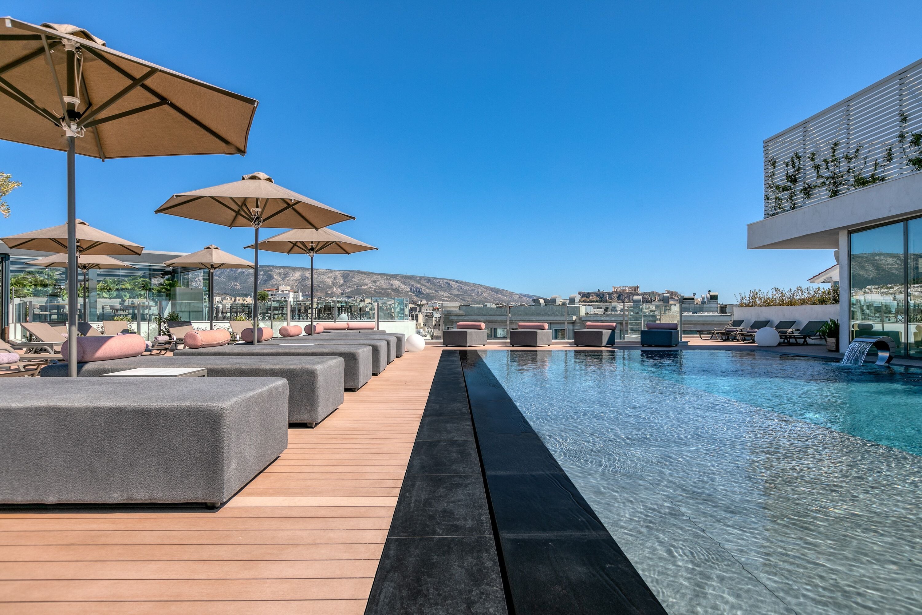 A rooftop pool area with lounge chairs and umbrellas, a wooden deck, and a clear view of the city and mountains in the background.