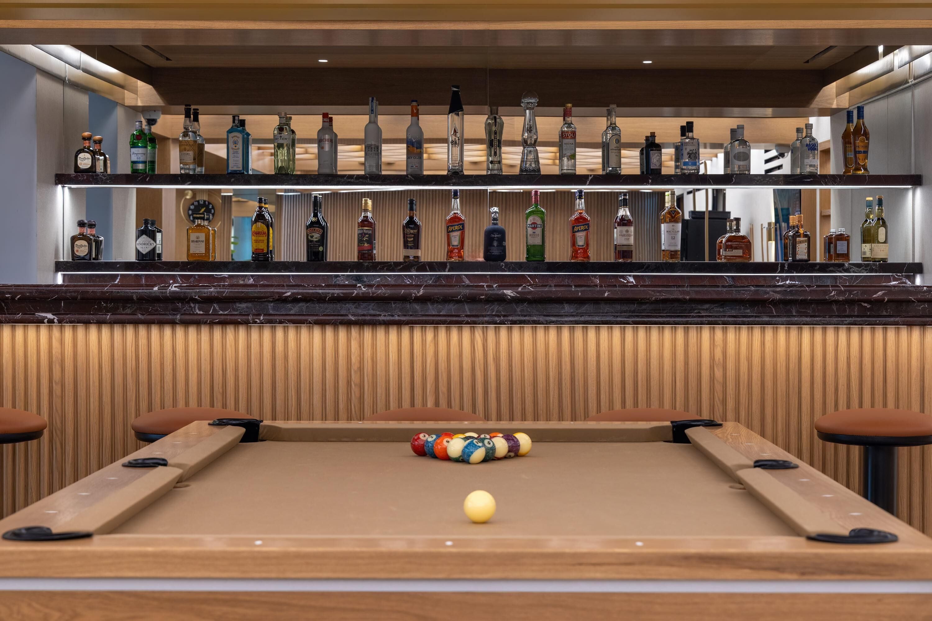 Mesa de bilhar com bola branca e uma bola listrada, área de bar com diversas garrafas de bebidas e parede de madeira com ripas verticais.