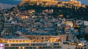 A cityscape with a prominent ancient Greek temple on a hill, modern buildings, and a waterfront area with a restaurant.