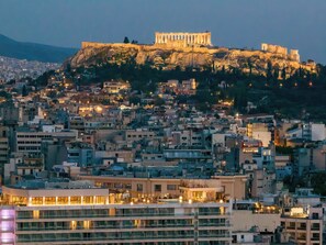 A cityscape with a prominent ancient Greek temple on a hill, modern buildings, and a waterfront area with a restaurant.