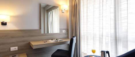 A hotel room with a wooden desk, a chair, a round table with breakfast items, a large mirror, and a window with curtains.