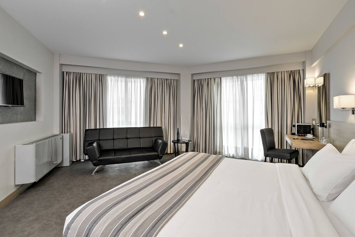 A hotel room with a large bed, a sofa, a desk with a chair, a TV mounted on the wall, and a window with curtains.
