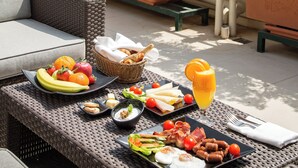 An outdoor dining area with wicker furniture, a variety of food and drinks, and a view of the sea and mountains in the background.