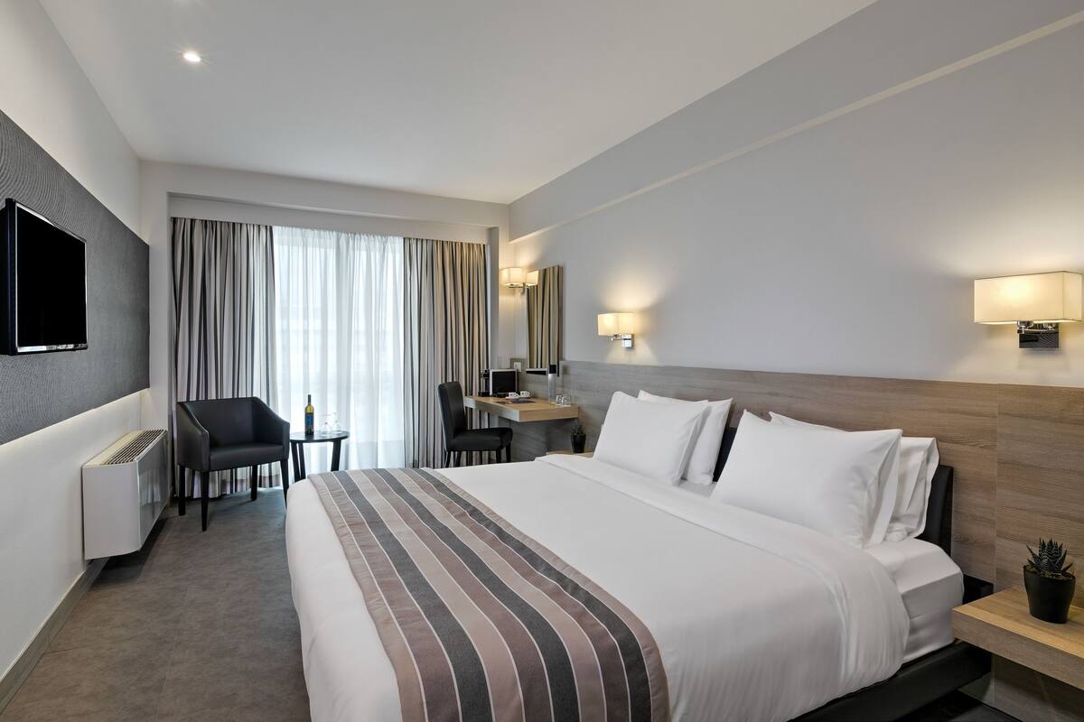 A hotel room with a large bed, a desk with a chair, a TV mounted on the wall, and a window with curtains.