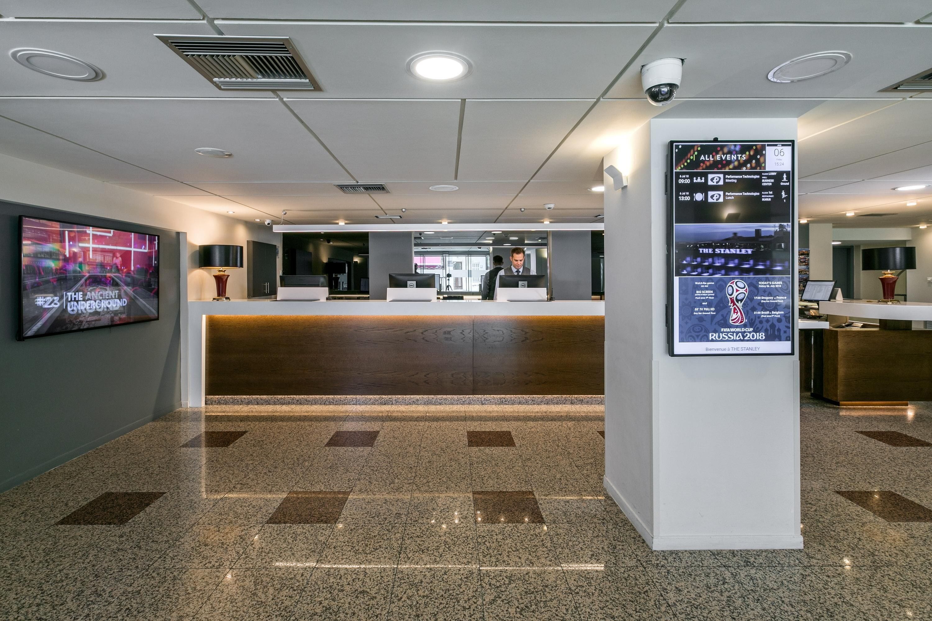 A modern hotel lobby with a reception desk, a digital information board, and a large screen displaying a movie.