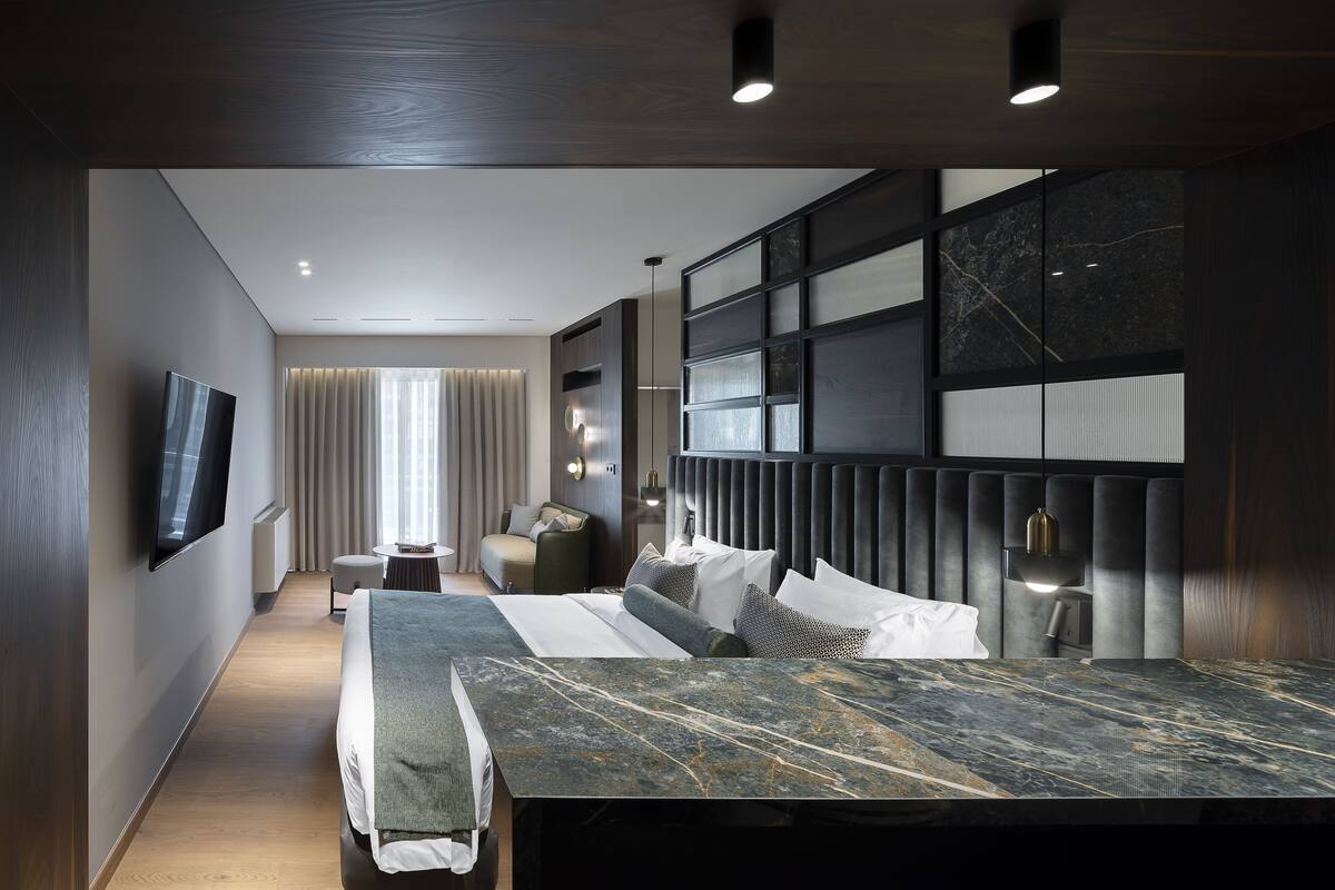 A modern hotel room with a large bed, a TV mounted on the wall, a seating area with a sofa and armchairs, and a large window with curtains.
