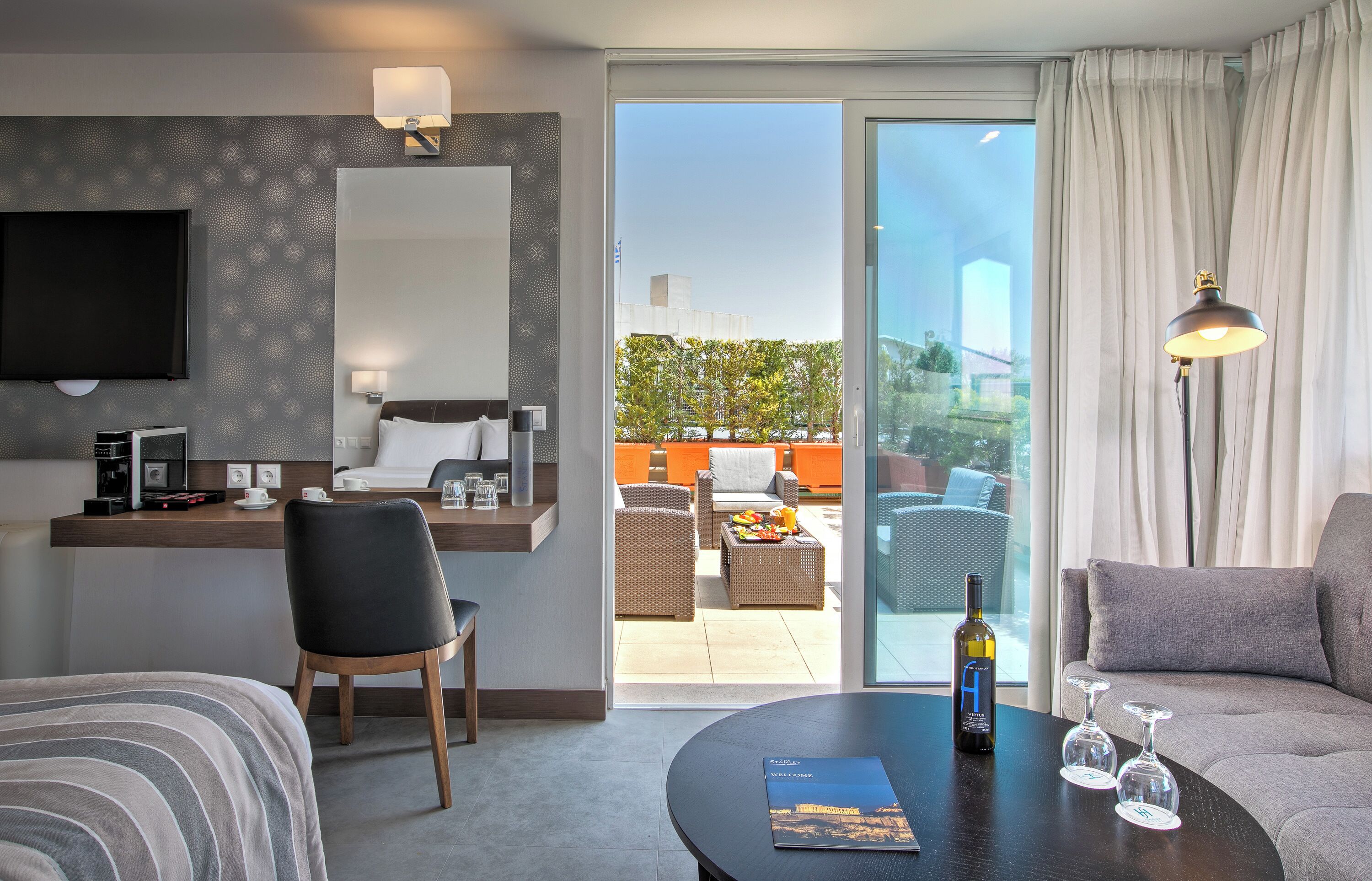 A hotel room with a flat-screen TV, a desk with a coffee maker, a chair, a sofa, a dining table with a wine bottle and glasses, and a sliding glass door to an outdoor seating area.