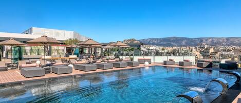 A rooftop infinity pool with lounge chairs and umbrellas, overlooking a cityscape and mountains.