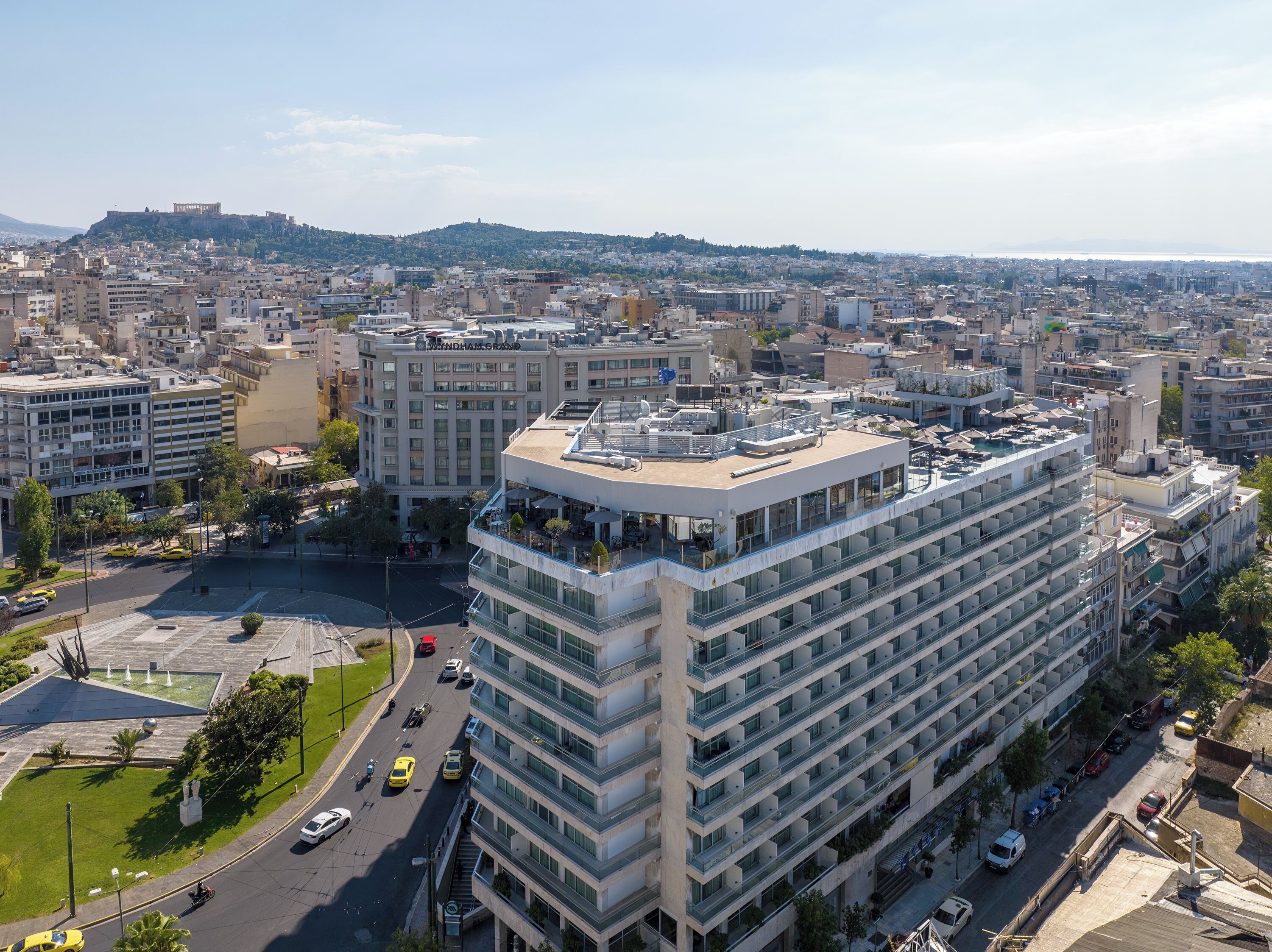 A multi-story hotel with a rooftop terrace, surrounded by a cityscape with various buildings and a distant hill.