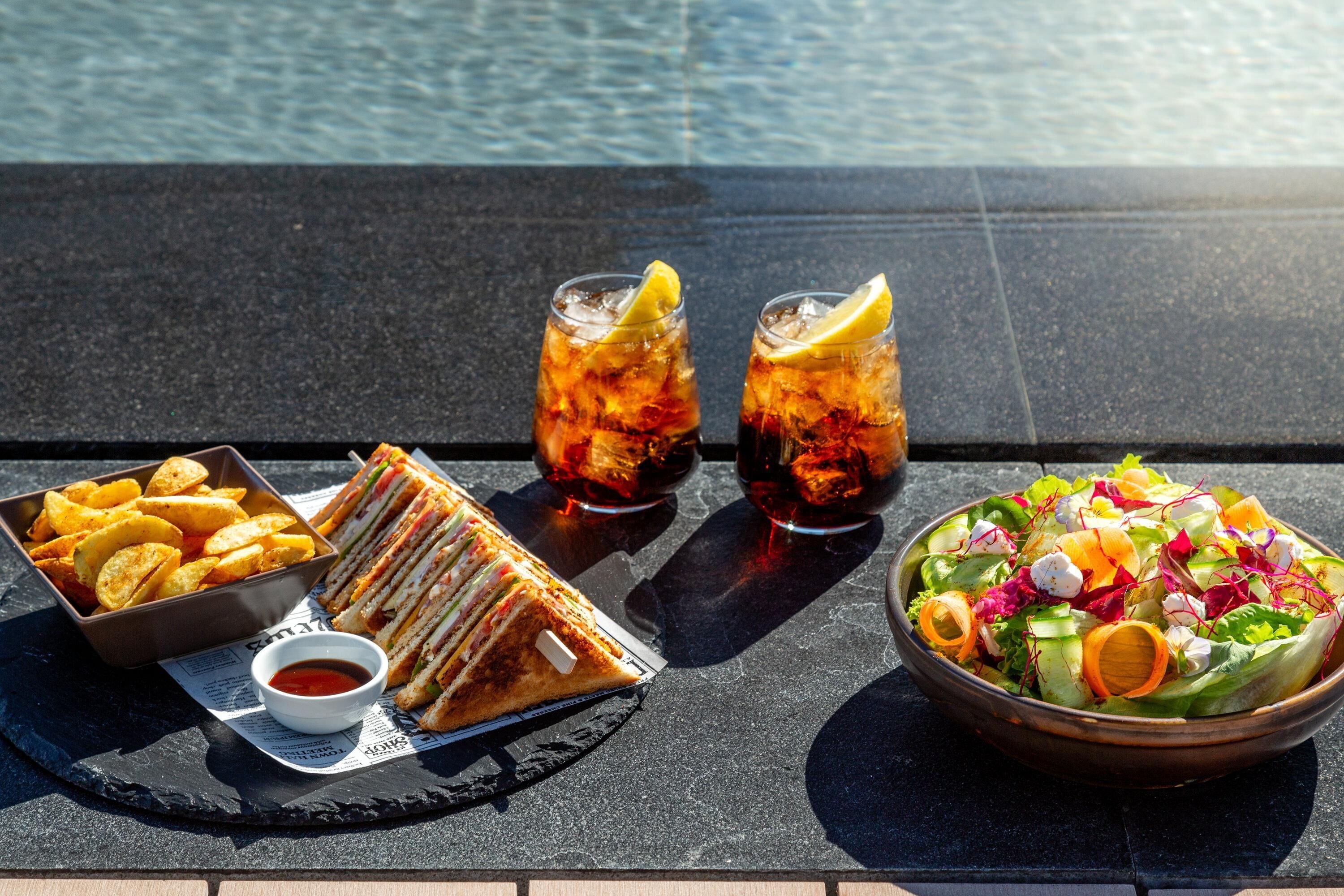 A table with a sandwich, chips, two drinks, a salad, and a small bowl of sauce.
