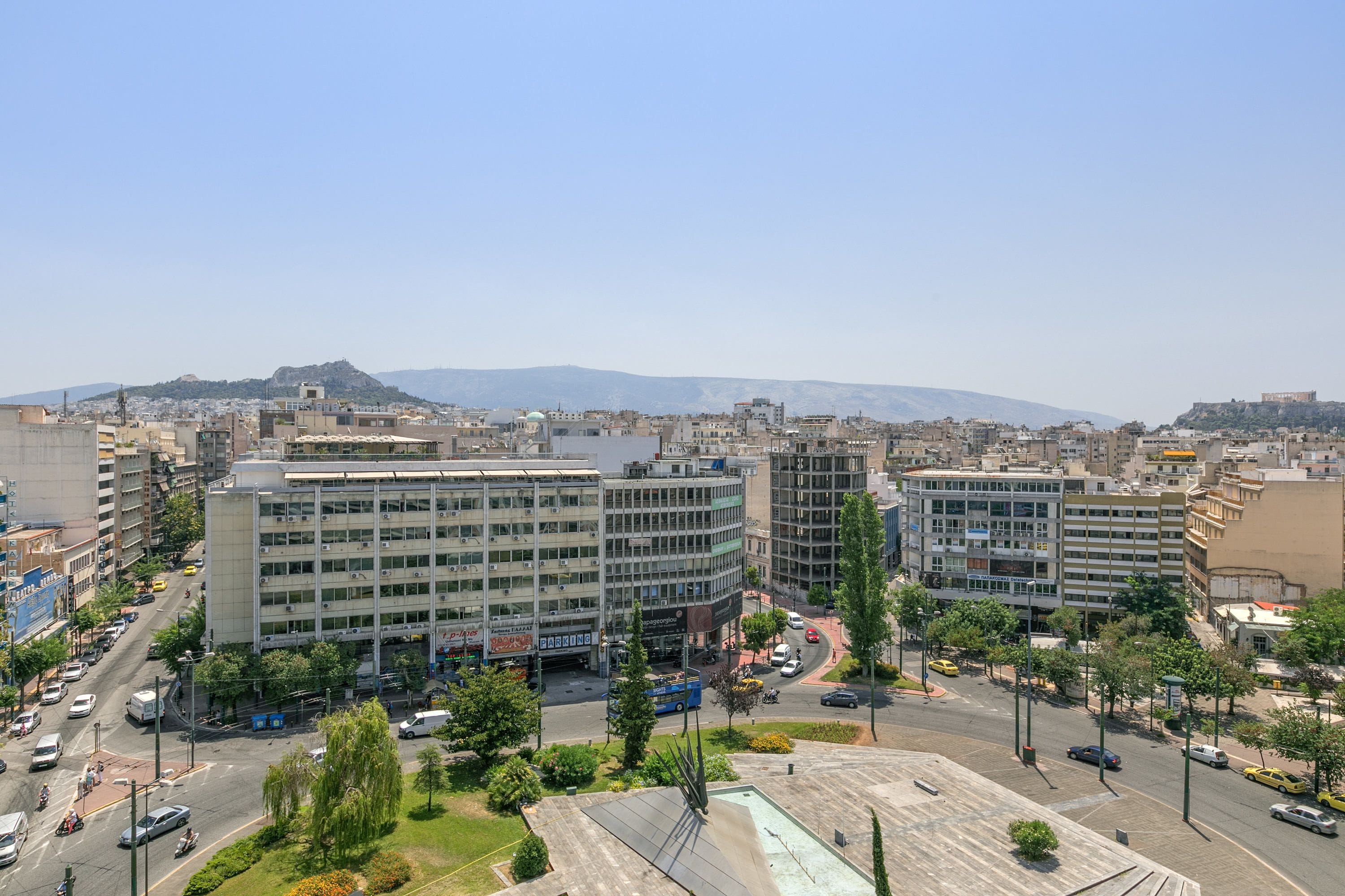 A cityscape with modern buildings, a central roundabout, and a distant mountain range.