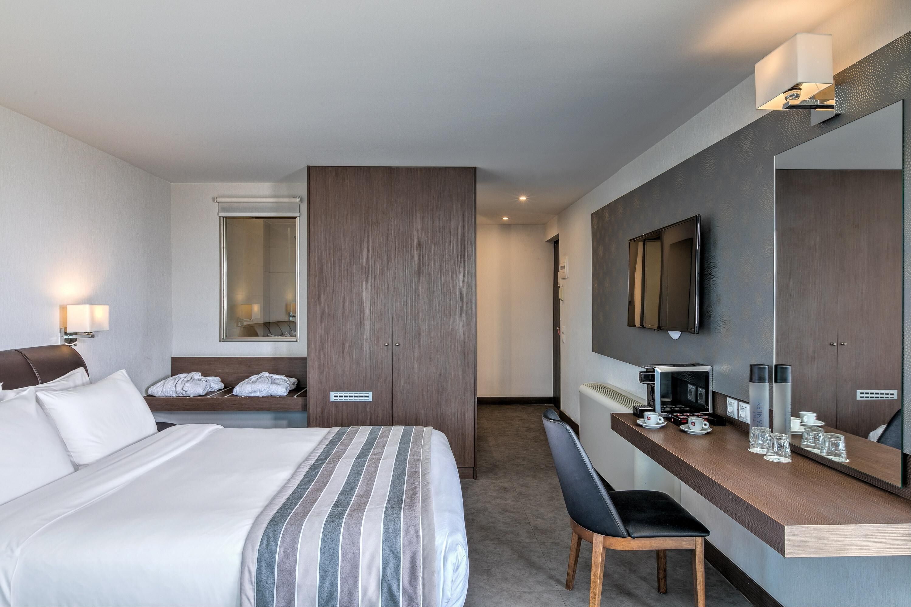 Quarto de hotel com cama, escrivaninha com cafeteira, televisão e espelho.