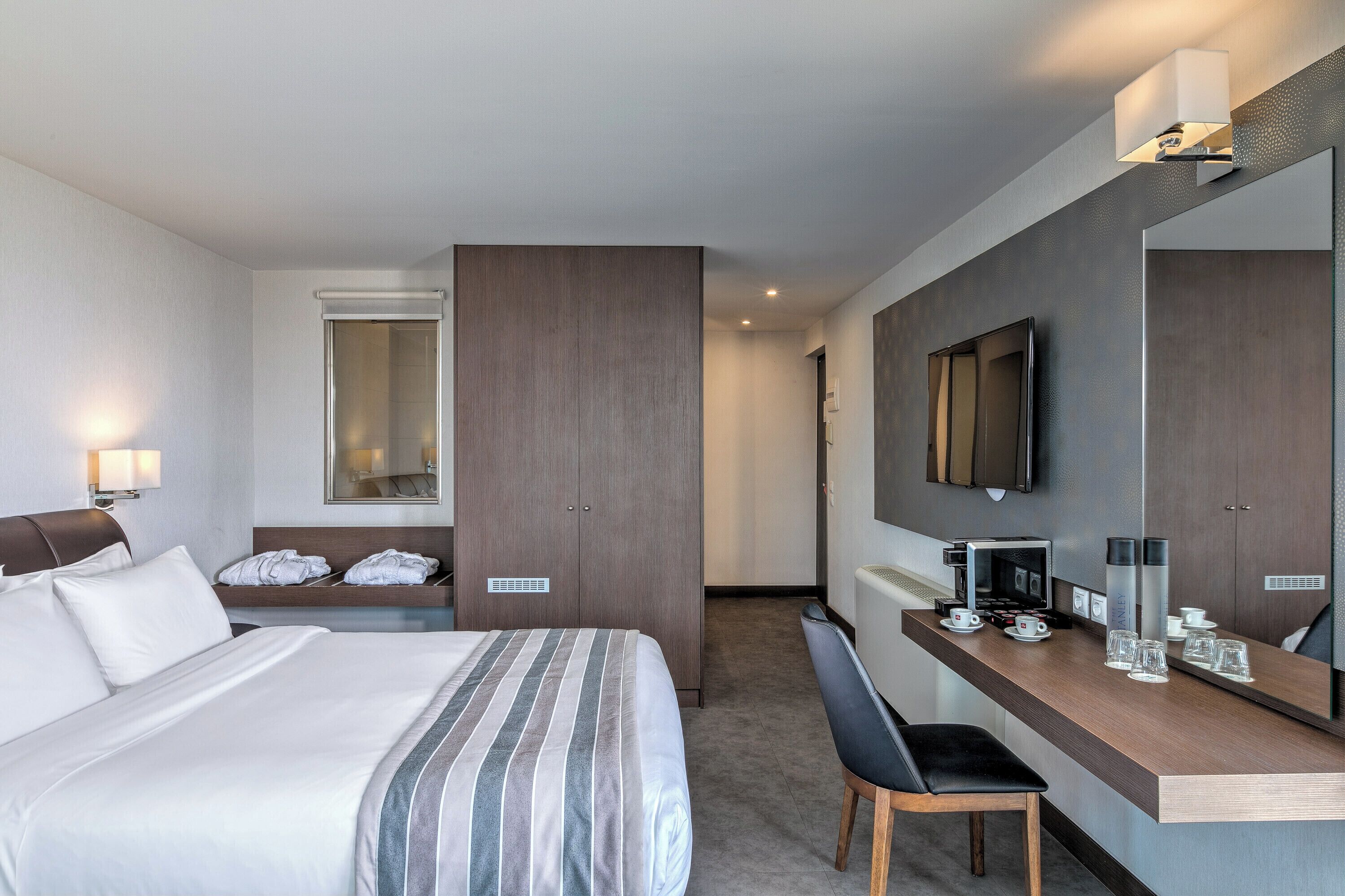 A hotel room with a bed, a desk with a coffee maker, a chair, a TV, and a mirror.
