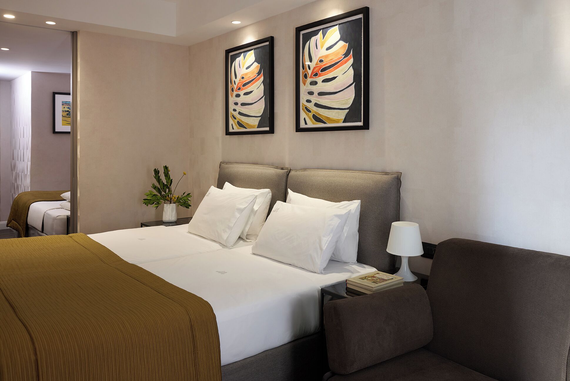 A hotel room with a large bed, two pillows, a bedside table with a lamp, and two framed artworks on the wall.