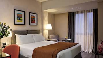 A hotel room with a large bed, a desk, a chair, a lamp, and two framed pictures on the wall.
