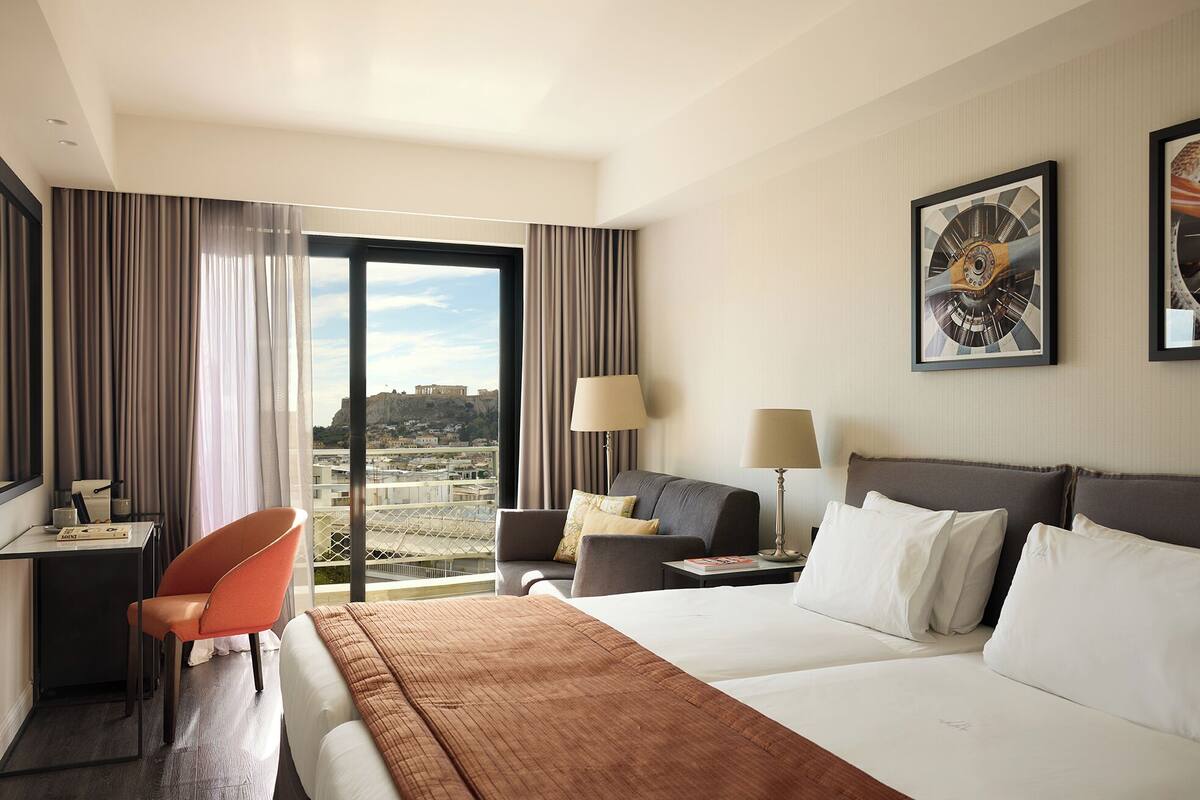 A hotel room with a bed, a desk, a chair, a sofa, a lamp, and a view of a cityscape.