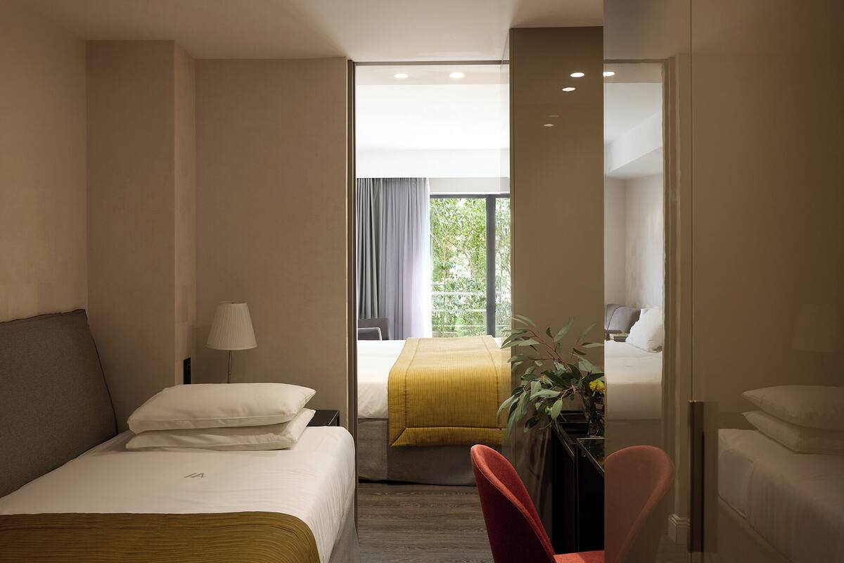 A hotel room with a bed, a desk, a chair, a lamp, and a view of a balcony with greenery.